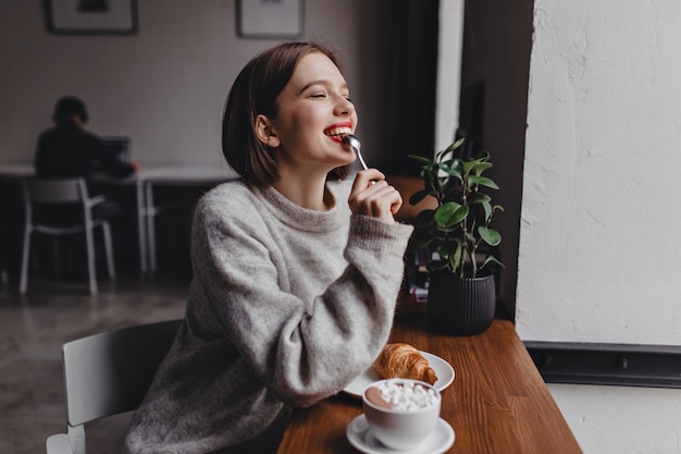 Short-haired lady in gray sweater bites teaspoon. Portrait of girl with red lips sitting in cafe and enjoying croissant and cappuccino.