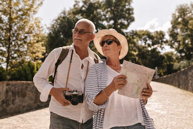 Short haired lady in hat, cool sunglasses and striped blue clothes holding map and posing with man in glasses and white shirt with camera in park.