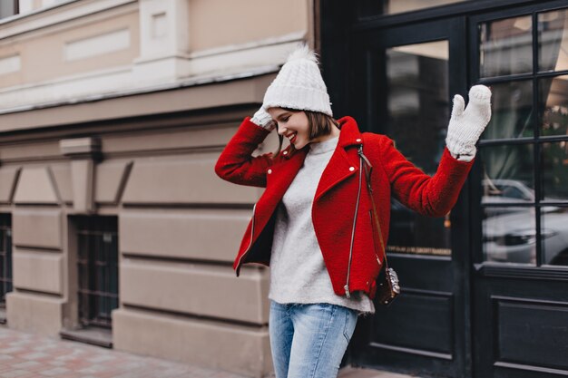 Short-haired lady in white hat and mittens is laughing. Portrait of girl with red lipstick dressed in bright jacket and jeans on background of shopwindow.