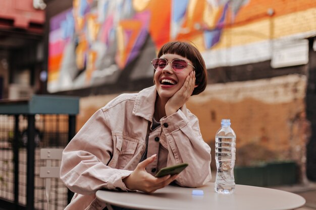 Short-haired teen in bright glasses and light clothes holding phone and laughing at street