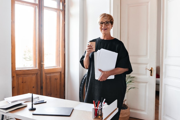 Short haired woman in black outfit holding coffee cup and paper sheets