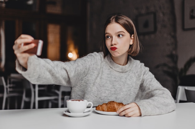 Short-haired woman in cashmere sweatshirt ordered croissant and cappuccino at cafe and takes selfie.