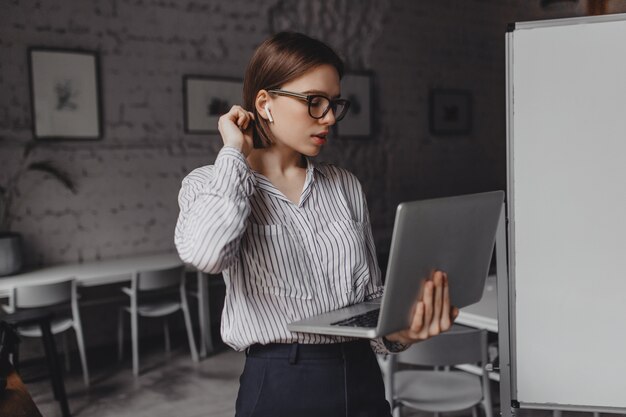 Short-haired woman in headphones speaks by video call and holds laptop while working in spacious office.