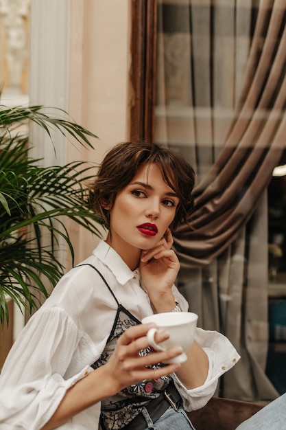 Short-haired woman in long sleeve shirt with red lips holding cup of coffee in restaurant. woman with brunette hairstyle poses in cafe.