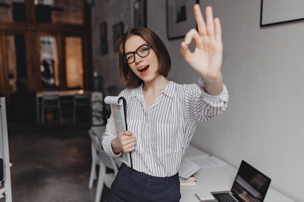 Short-haired woman in striped blouse shows ok sign. portrait of female worker in glasses winking and leaning on table with laptop.