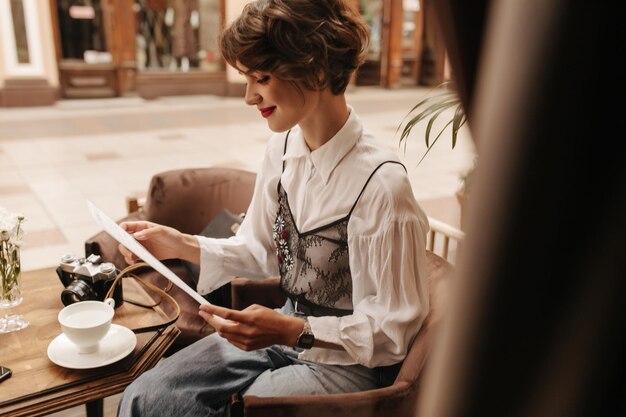 밝은 입술을 가진 짧은 머리 소녀는 미소를 지으며 카페에 앉아 있습니다. 현대적인 셔츠와 청바지를 입은 젊은 여성이 레스토랑에서 메뉴를 읽고 있습니다.