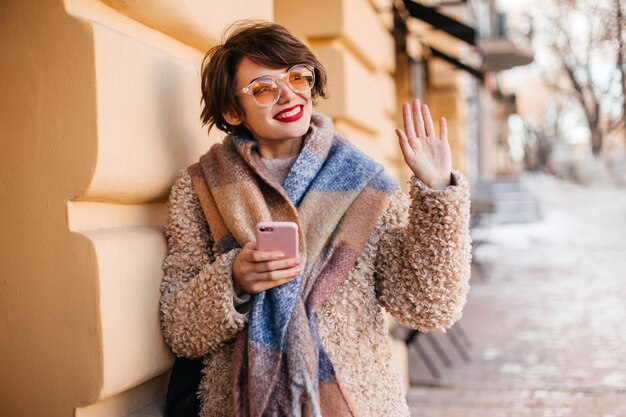 Shorthaired woman with scarf waving hand Outdoor shot of cheerful girl holding smartphone on street