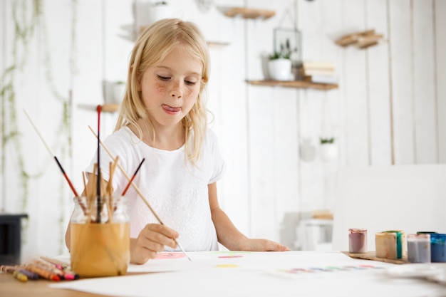 Shot of adorable blonde girl with freckles biting her tongue because of inspiration while painting. Girl with blond hair sitting at the room filled with morning light and wearing white clothes.