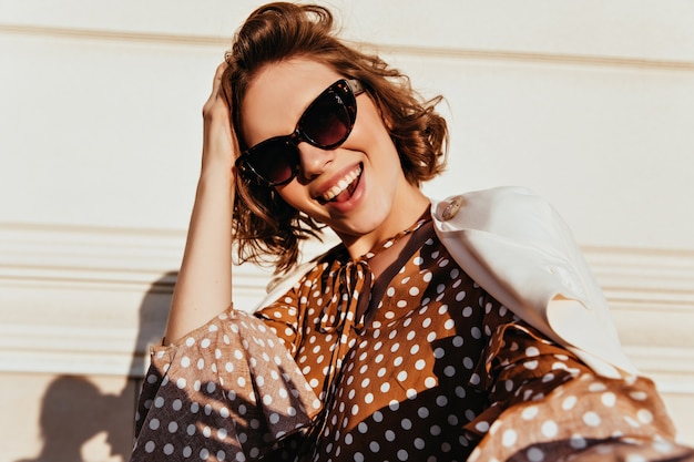 shot of adorable pleased woman in black sunglasses. Outdoor shot of refined curly lady expressing happiness.