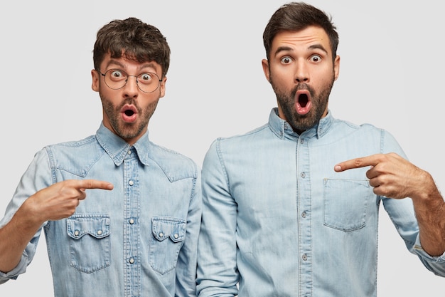 shot of amazed bearded guys with stupefied expression point at each other with index fingers, wear denim shirts, opens mouthes widely, stand against white wall. Friendship concept