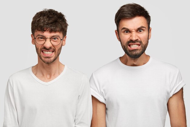 shot of angry furious bearded men colleagues clench teeth in annoyance, feel irritation as recieve much work and duties from boss, dressed in casual white t-shirts. Negative human emotions