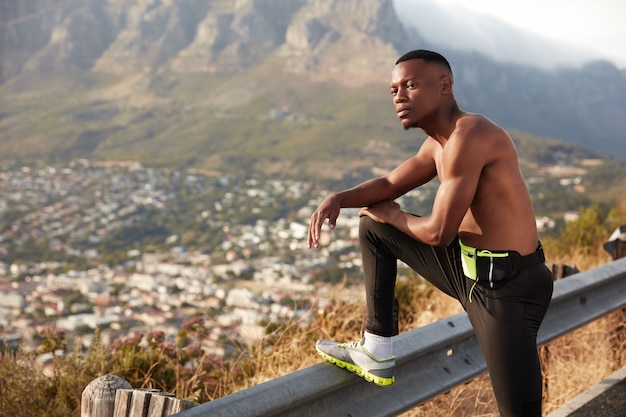 Shot of athlete man with dark healthy skin, has rest after physical exercises, keeps legs raised on traffic sign, has thoughtful expression, poses in moutains enjoys sport in open air. Jogging concept