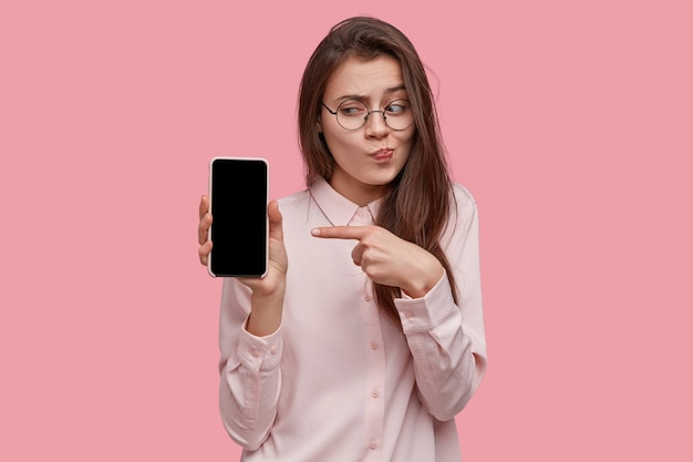 Shot of attractive dark haired female keeps modern cell phone in hand with mock up screen, advertises new gadget of her favourite company