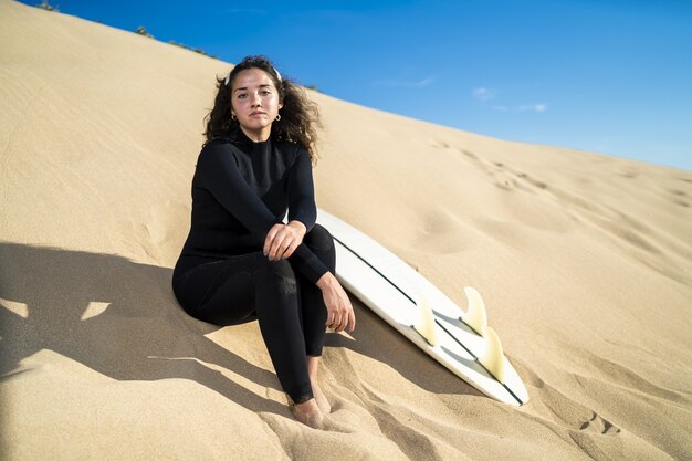 shot of an attractive female sitting on a sandy hill with a surfboard on the side