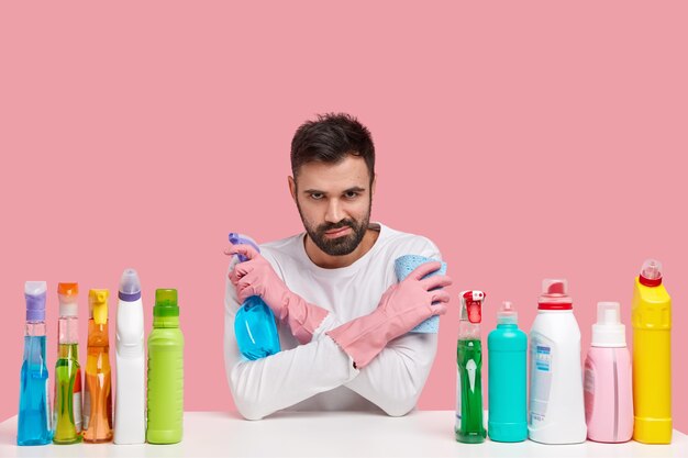 Shot of attractive man crosses hands over hands, carries washing spray and rag, looks with gloomy expression, wears casual clothes