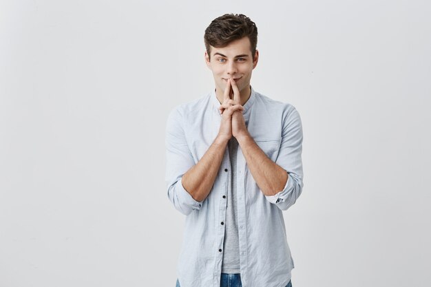 Shot of attractive young caucasian man dressed casually looking  with cunning thoughtful expression, keeping index fingers on his face, trying to recollect something or having great idea