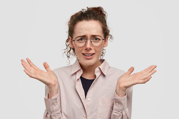 shot of attractive young female with hesitant expression, shruggs shoulders and keeps palms raised, has clueless look, isolated over white wall. Puzzlement and doubt concept.