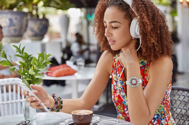 Shot of beautiful concentrated female has afro hairstyle searches favourite song in playlist, enjoys loud music in headphone while sits at outdoor cafeteria
