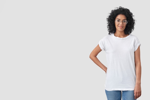 shot of beautiful female journalist ready to take interview, keeps one hand on waist, has pleased confident expression wears white oversized t-shirt, jeans and spectacles, poses over blank wall