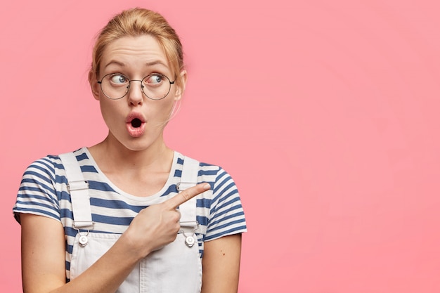 Shot of beautiful shocked female with light hair and blue eyes, indicates at blank copy space for your advertisment, sees something unexpected away