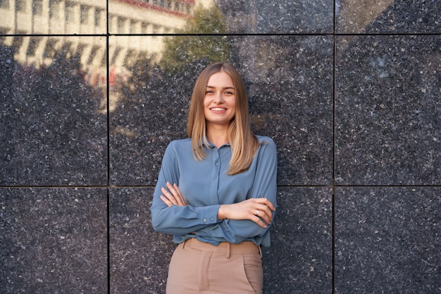 Shot of beautiful young businesswoman wearing blue chiffon shirt while standing with folded arms on gray marble wall