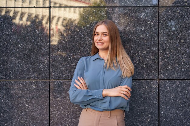 Shot of beautiful young businesswoman wearing blue chiffon shirt while standing with folded arms on gray marble wall