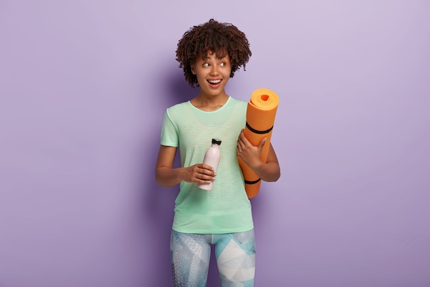 shot of cheerful dark skinned girl holds fitness mat and bottle of fresh water, drinks during exhausted exercising looks right dressed in active wear poses indoor. Motivation, healthy lifestyle