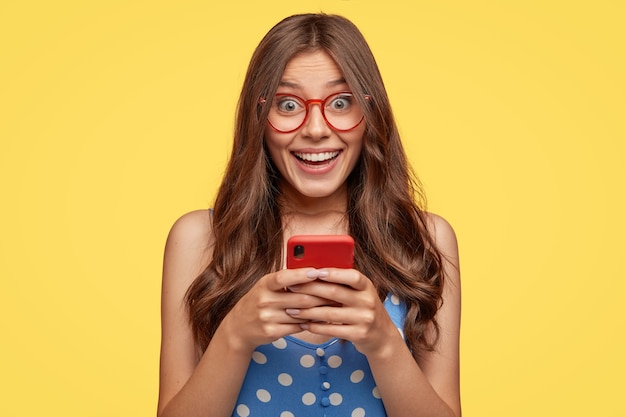 Shot of cheerful female model wears glasses, holds cell phone, takes pictures, talks via video message, has nice friendly talk, dressed in fashionable outfit, isolated over yellow wall
