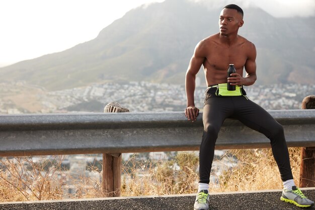 Shot of dark skinned man has intensive preparation to sport competitions, holds bottle of water, looks thoughtfully aside, wears sneakers and leggings, full of energy.