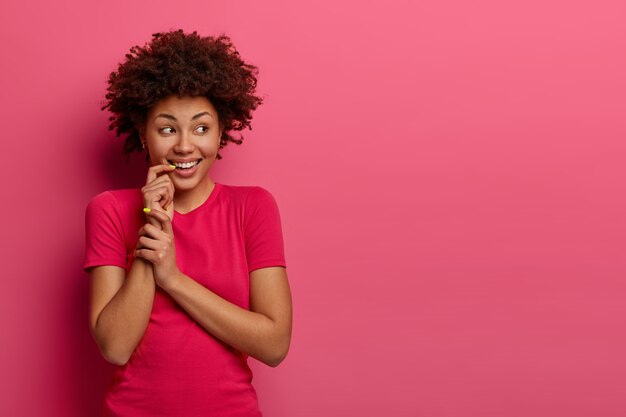 shot of dark skinned woman giggles positively, looks aside, keeps hand near mouth, feels joyful, notices something appealing, being amused, dressed casually, isolated on pink wall, free space