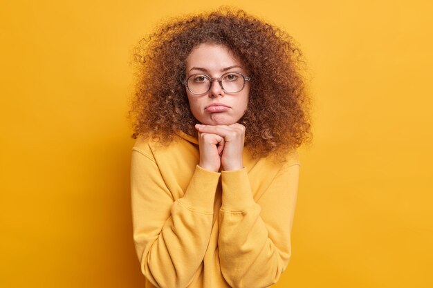  shot of disappointed sad European woman with curly hair feels upset keeps hands under chin purses lips dressed in sweatshirt isolated over yellow wall feels troublesome and bored