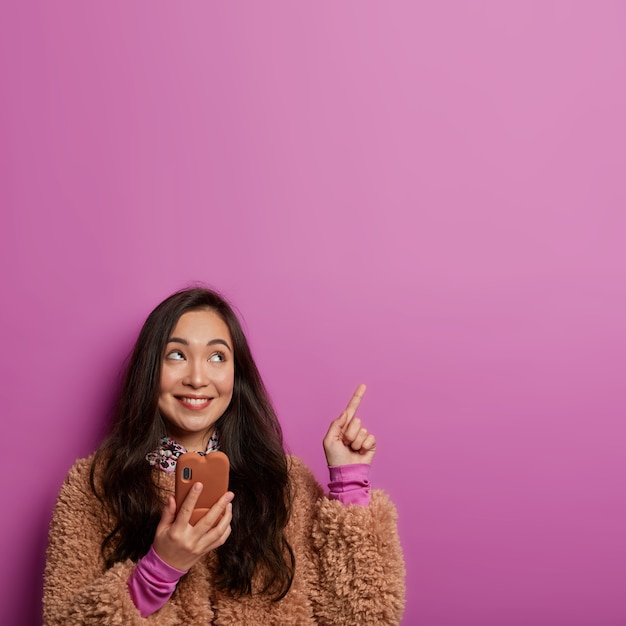 Shot of dreamy young woman with toothy smile, dark hair, has interesting idea, recommends something on blank space, uses mobile phone