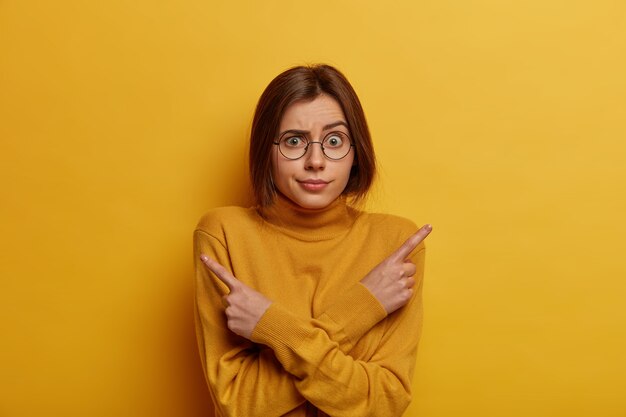 shot of embarrassed unaware woman points sideways, thinks what to choose, raises eyebrows, indicates right and left, dressed in yellow outfit, stands indecisive indoor. Difficult choice