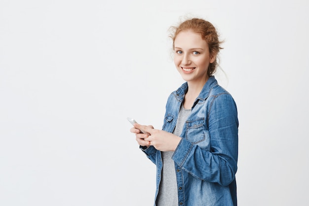 shot of emotive young redhead girl with cute smile, standing half-turned with combed hair, holding smartphone while glancing at camera, wearing denim shirt