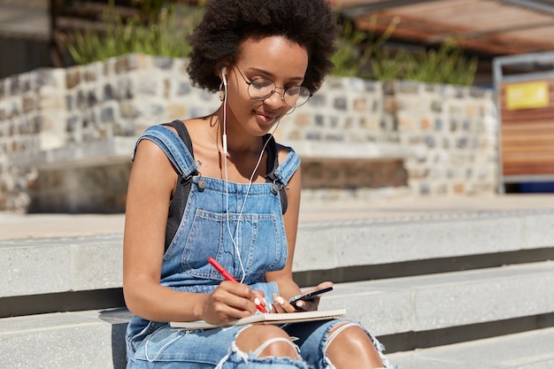 Free photo shot of female student listens audio book with earphones and mobile phone, writes some records and details in diary, poses at stairs outdoor, prepares for seminar, uses internet and technology.