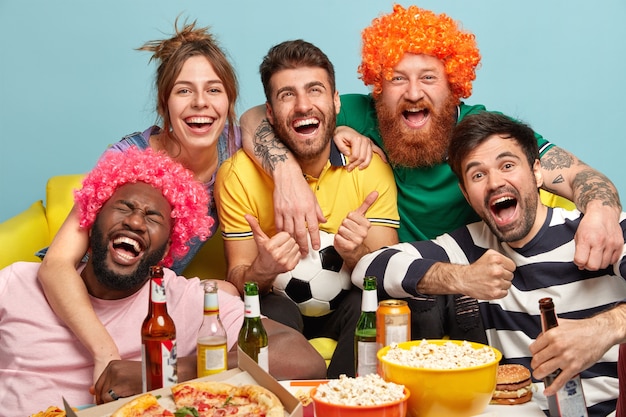 Shot of friendly companions embrace and smile happily, cheer with winning favorite team, have nice time together watching exciting football game, drink beer and eat fast food. Funny fans support