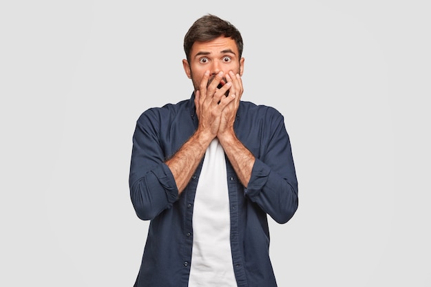 Free photo shot of frightened caucasian male covers mouth with both hands, stares with bugged eyes, dressed in fashionable outfit, reacts on something, isolated over white wall. emotions concept