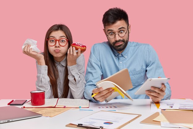 Shot of funny brunette woman eats delicious doughnut, has full mouth while her partner realizes shocking news, stares at notepad, holds touchpad