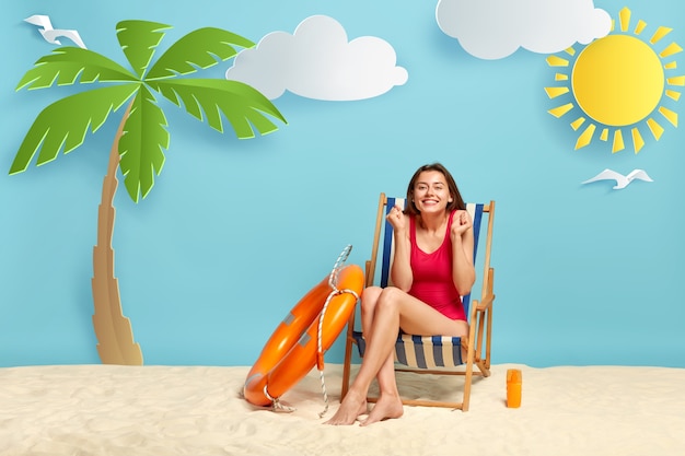 Free photo shot of glad female vacationist in red bikini, clenches fists, enjoys recreation time, poses at beach chair