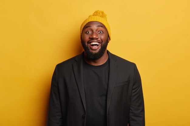 Shot of glad plump dark skinned man laughs positively, smiles joyfully, wears black elegant clothes and bright headgear, stands against yellow background, feels happiness