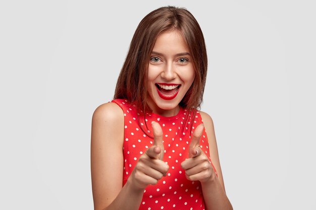 shot of glad young female with positive expression points with both index fingers, isolated over white wall. Cheerful woman shows gun gesture, greets friend, approves idea