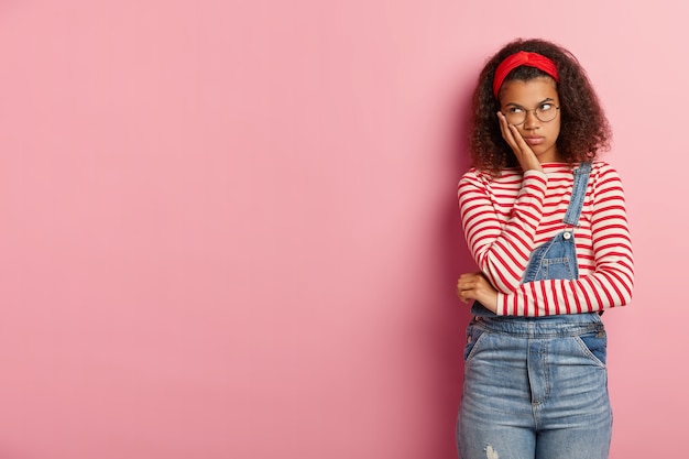 Shot of gloomy unhappy teenage girl posing in overalls with curly hair