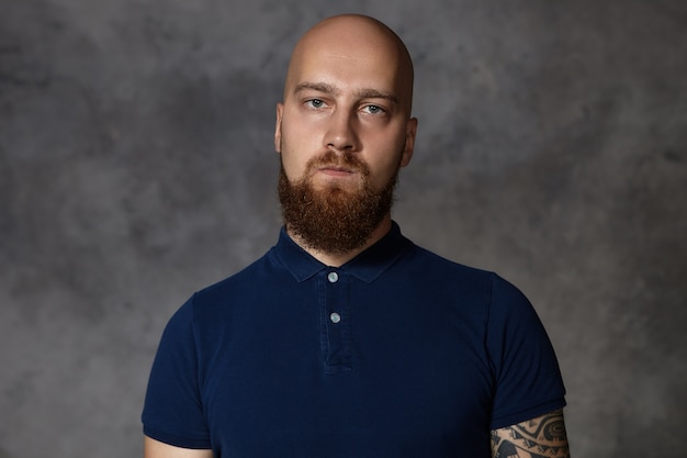 Free photo shot of gloomy young bald male with bushy beard being dissatisfied with unfair results of competition, pursuing lips, trying to hide his anger,