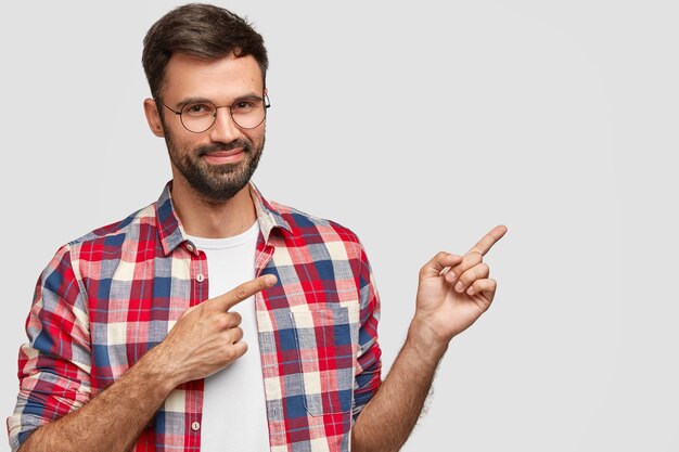 shot of good looking man with satisfied expression, has dark stubble, indicates with fore fingers aside