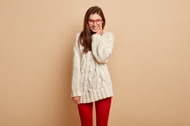 shot of good looking young woman smiles and poses indoor, has feeling of happiness, hears positive news, dressed in white jumper and red tights, shows perfect teeth, enjoys nice day off