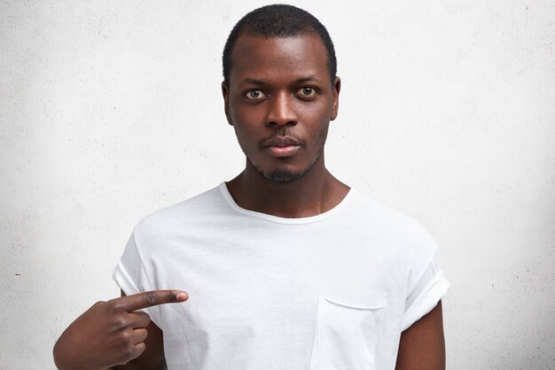 Shot of handsome serious young African male with confident expression, indicates with index finger at t shirt for your logo or advertising content.