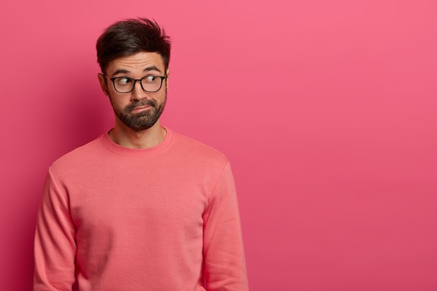 Free photo shot of handsome thoughtful man with dark thick beard concentrated aside on empty space of pink wall, notices interesting scene, has surprised expression, wears transparent glasses and jumper