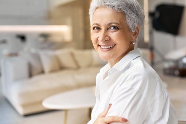 shot of happy 50 year old retired woman with freckles and gray hair posing on stylish interior background, wearing white shirt, smiling broadly at front