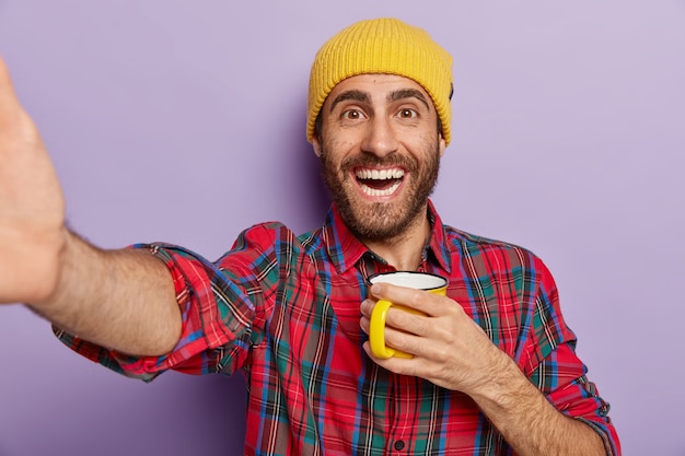 shot of happy Caucasian man takes selfie indoor, holds mug with coffee or tea, enjoys break and free time, wears stylish yellow hat and plaid shirt isolated on purple wall. People and lifestyle