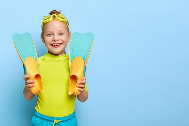 shot of happy ginger small girl shows new rubber flippers, wears swim goggles, dressed in summer clothes, enjoys learning swimming, has active rest, isolated on blue wall with blank space